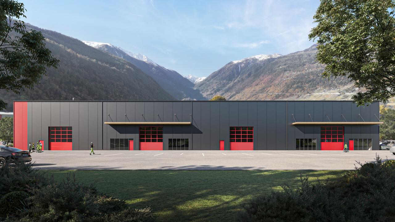 A warehouse-style building, mostly black with some red accents, has four large red garage doors in the center, large windows on the side, and a red pillar at the far left, situated on a large parking lot with mountain range in the background.