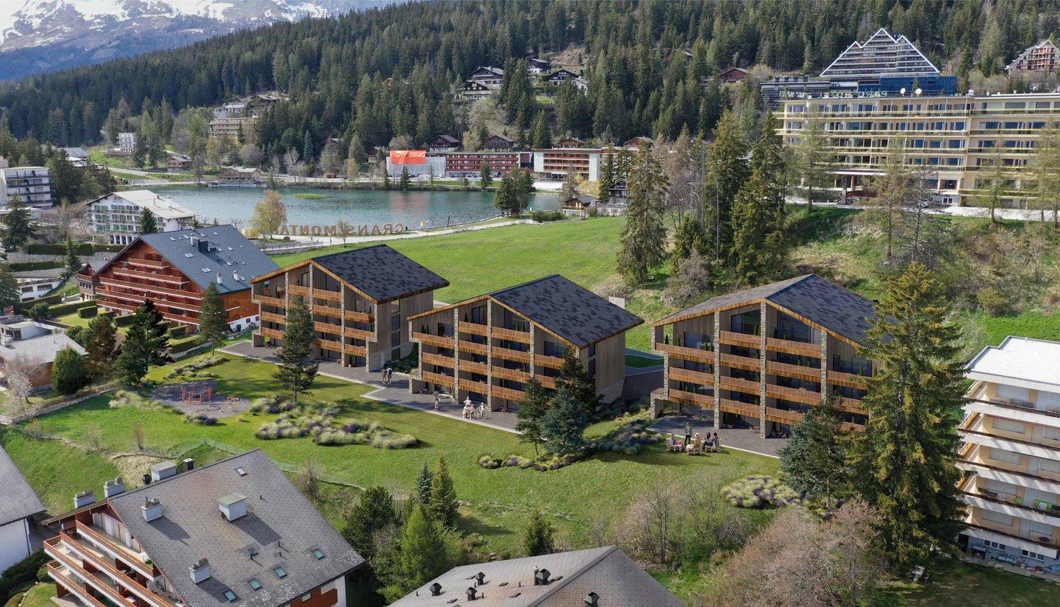 Aerial view of a resort complex with several chalet-style buildings set among trees and mountains. There is a lake and a mountain in the background. The area has lush greenery and is surrounded by a mountainous terrain.