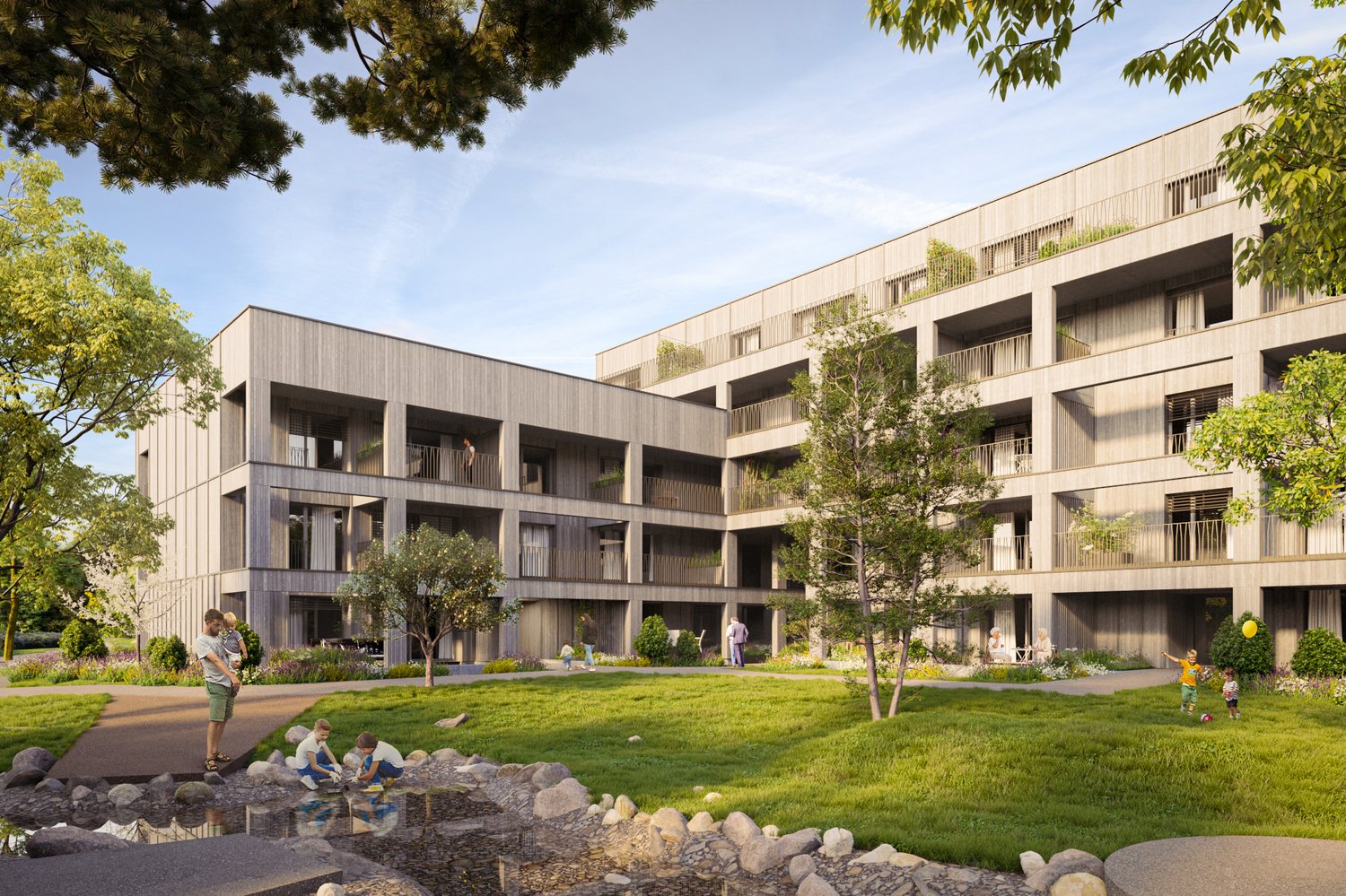 Multi-story apartment building with wooden exterior, balconies, and surrounded by trees and greenery. There is a grassy area with a small pond or water feature in the foreground, where children are playing.