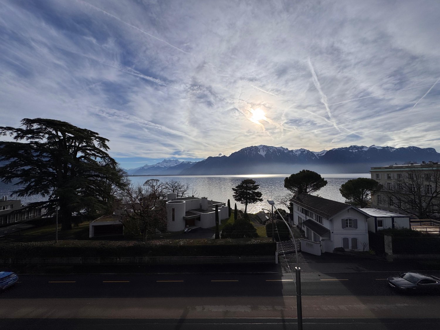 Wide view of a road, lake, mountains, white houses, road, trees, sky with clouds