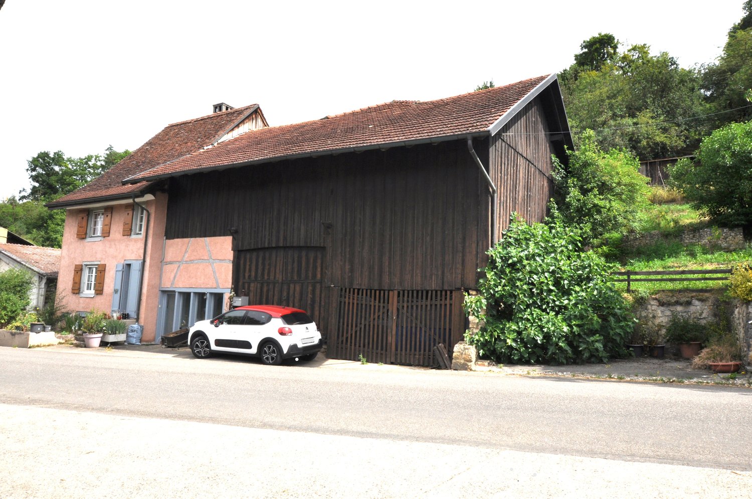 2-story house with a red tile roof, wooden siding, and a large garage attached to the side. The house is surrounded by trees and greenery.