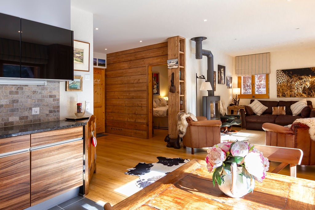 Open-concept living space with wooden walls and flooring, a stone fireplace, leather sofa, wooden dining table, and various decorative elements such as artwork and floral arrangements