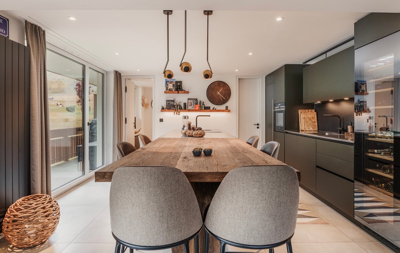 Dining room: wooden dining table, chairs, shelves with decorative items, wall clock, wooden flooring, kitchen area with sink and gas stove