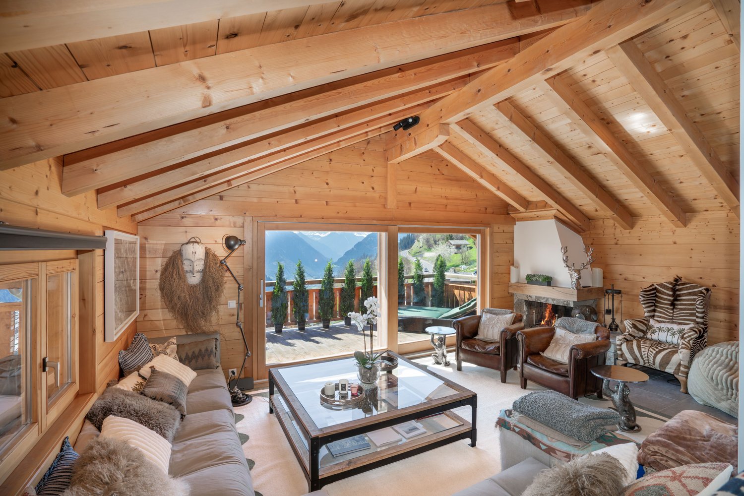 Living room with wooden walls, large windows, fireplace, glass table, couches, armchairs, and decorative items