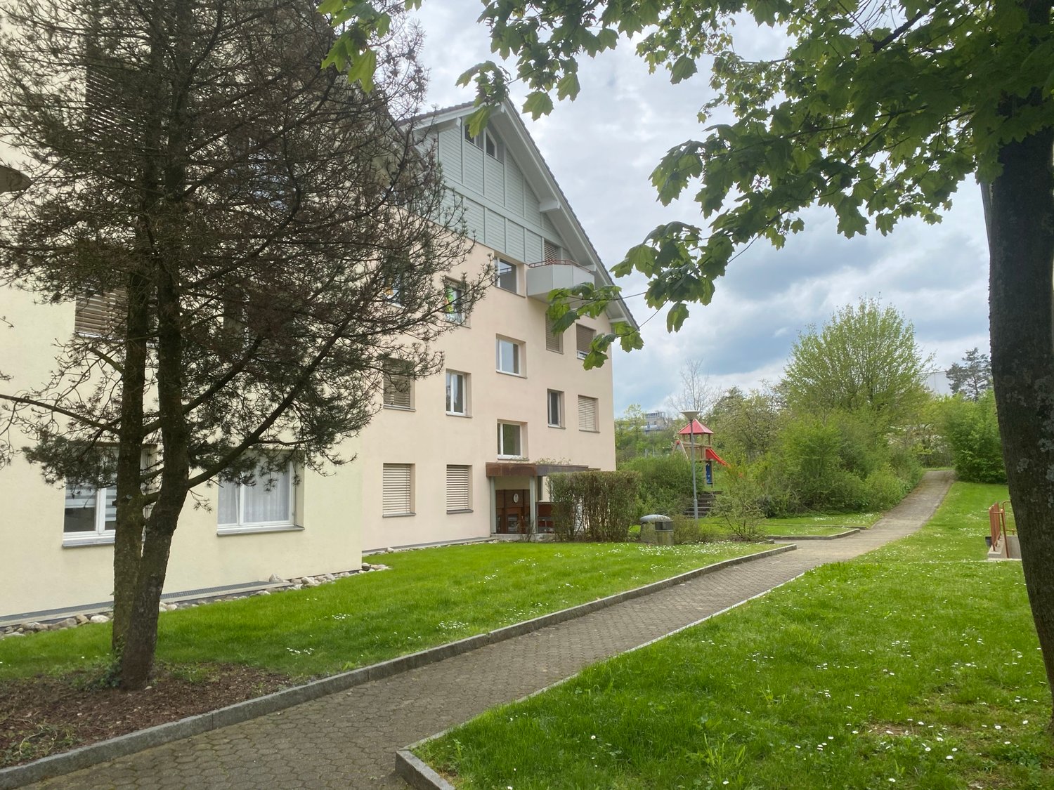 A multi-story building with beige walls and white windows, a small balcony on the second floor, and a pathway leading to the entrance. The building is surrounded by a well-maintained lawn and trees. In the distance, there is a playground with a red slide