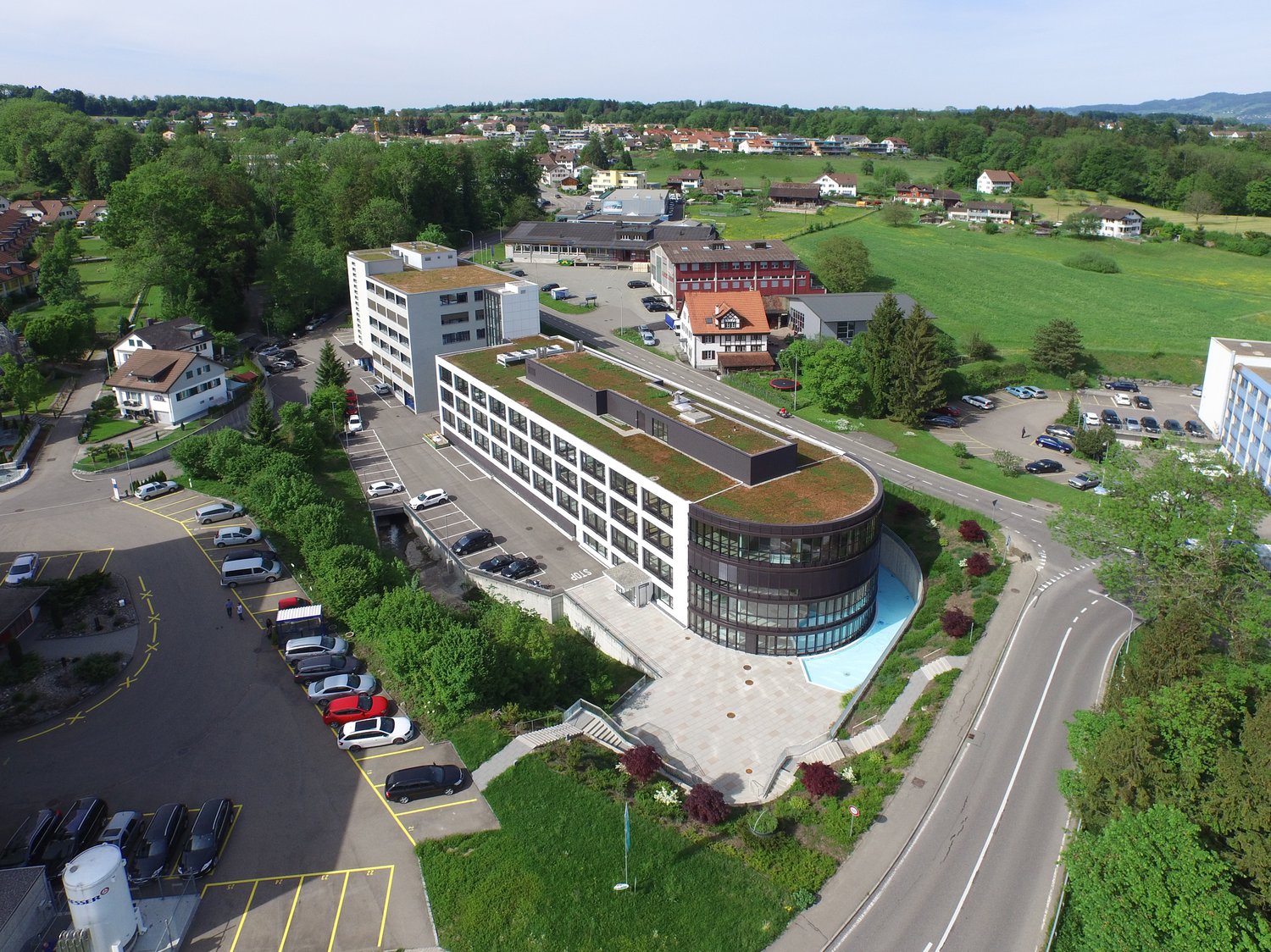 Multi-story office building with a curved glass facade, surrounded by a landscaped area with parking spaces. The building is situated in a green, hilly area with residential houses and other buildings in the background.