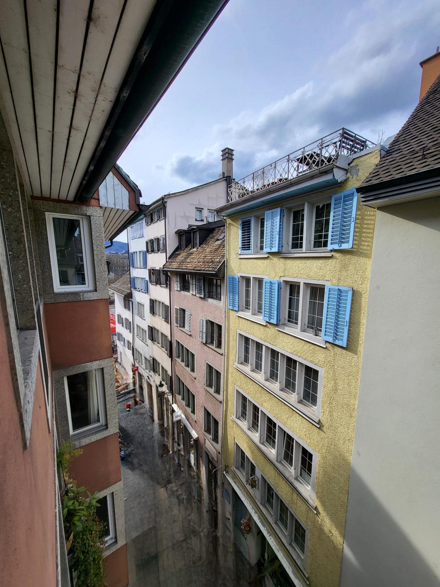 An aerial view of several buildings with various colors and blue shutters. The building in the center has a balcony with a metal railing.