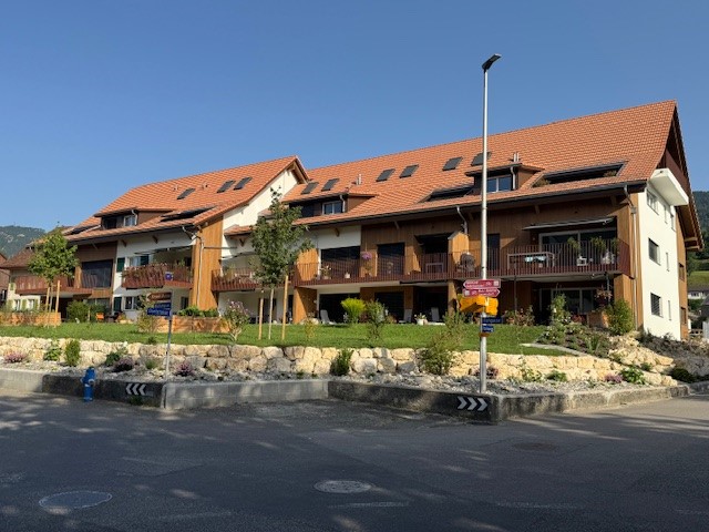 Apartment building with brown roof, balconies, large windows, stone wall, and trees in front