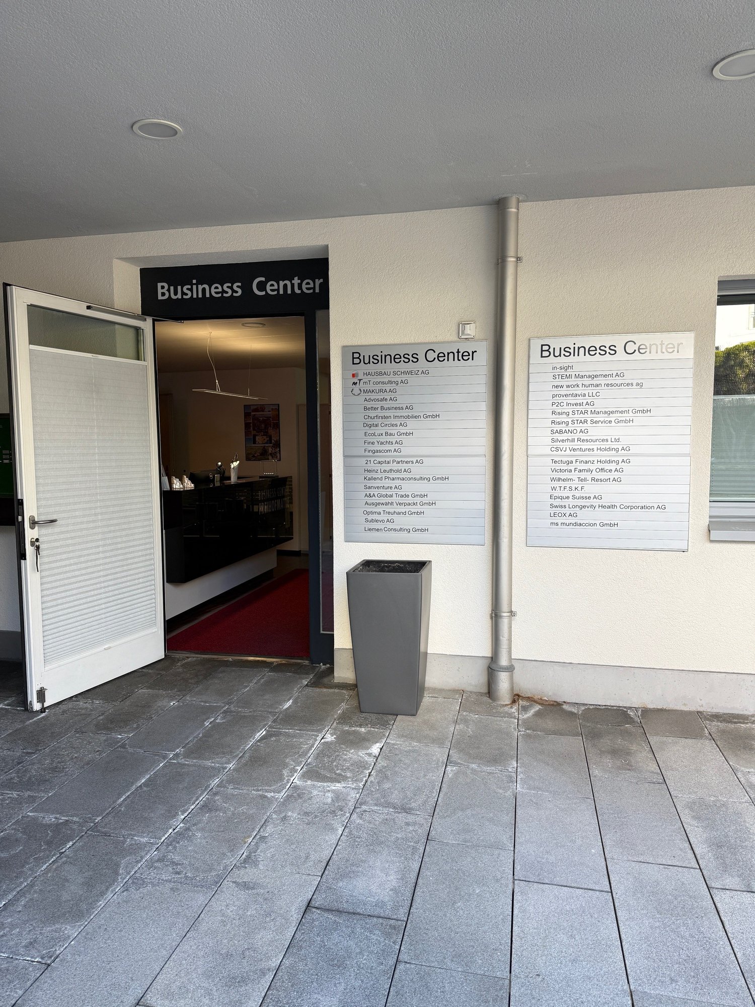 Entrance to a business center with an open door, a red carpet, a trash bin, and a signboard with the text 'Business Center'.