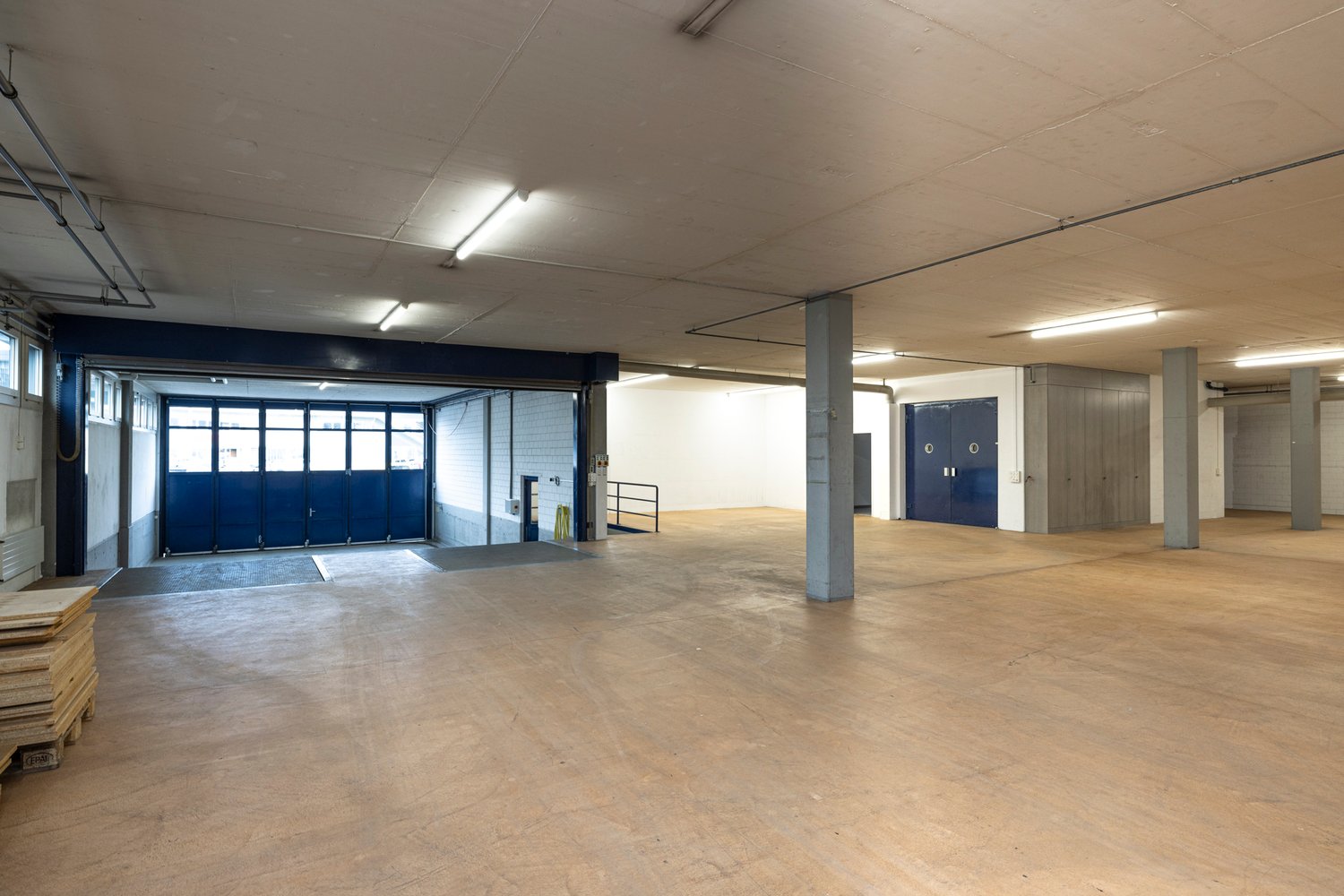 large open space, concrete floor, metal beams, large blue doors, storage boxes, stacks of wooden pallets