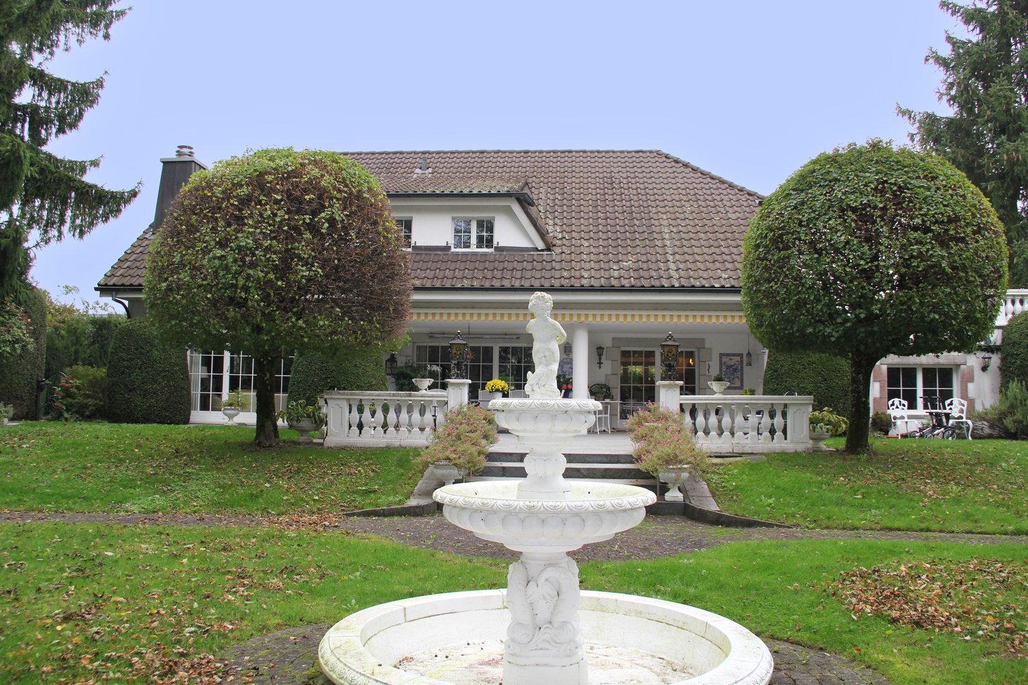 Large house with brown roof, white walls, large windows, white porch, garden with fountain, trees, and grass