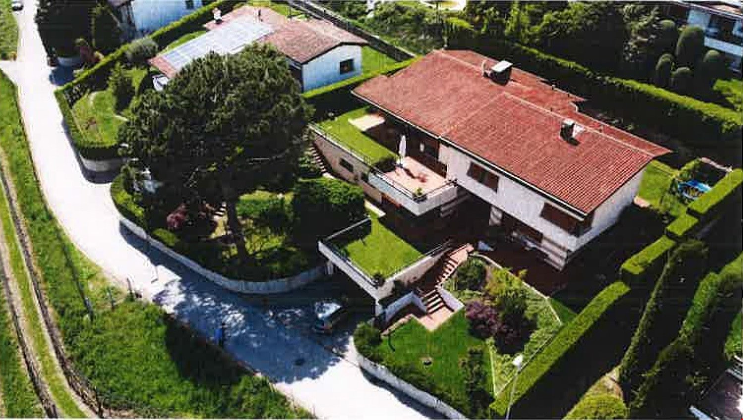 Large two-story house with red tile roof, surrounded by lush greenery and a swimming pool. The house has a balcony or terrace overlooking the landscaped garden.