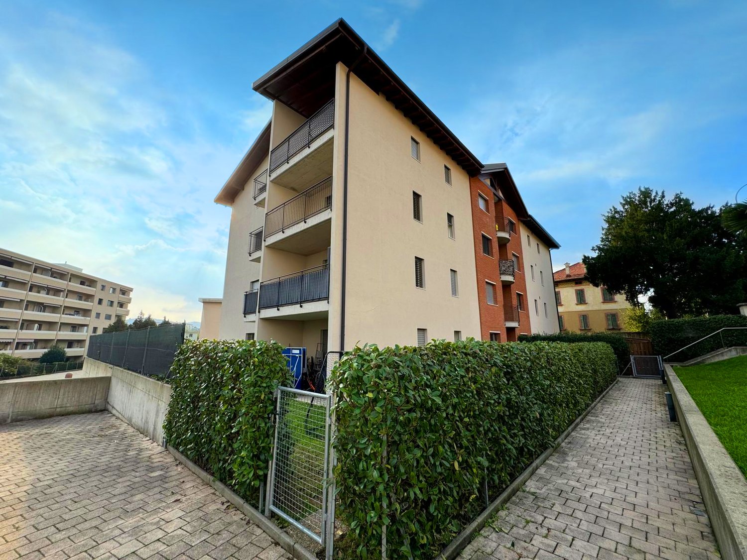 Multi-story apartment building with beige exterior, balconies, and a landscaped courtyard with hedges and a paved walkway.