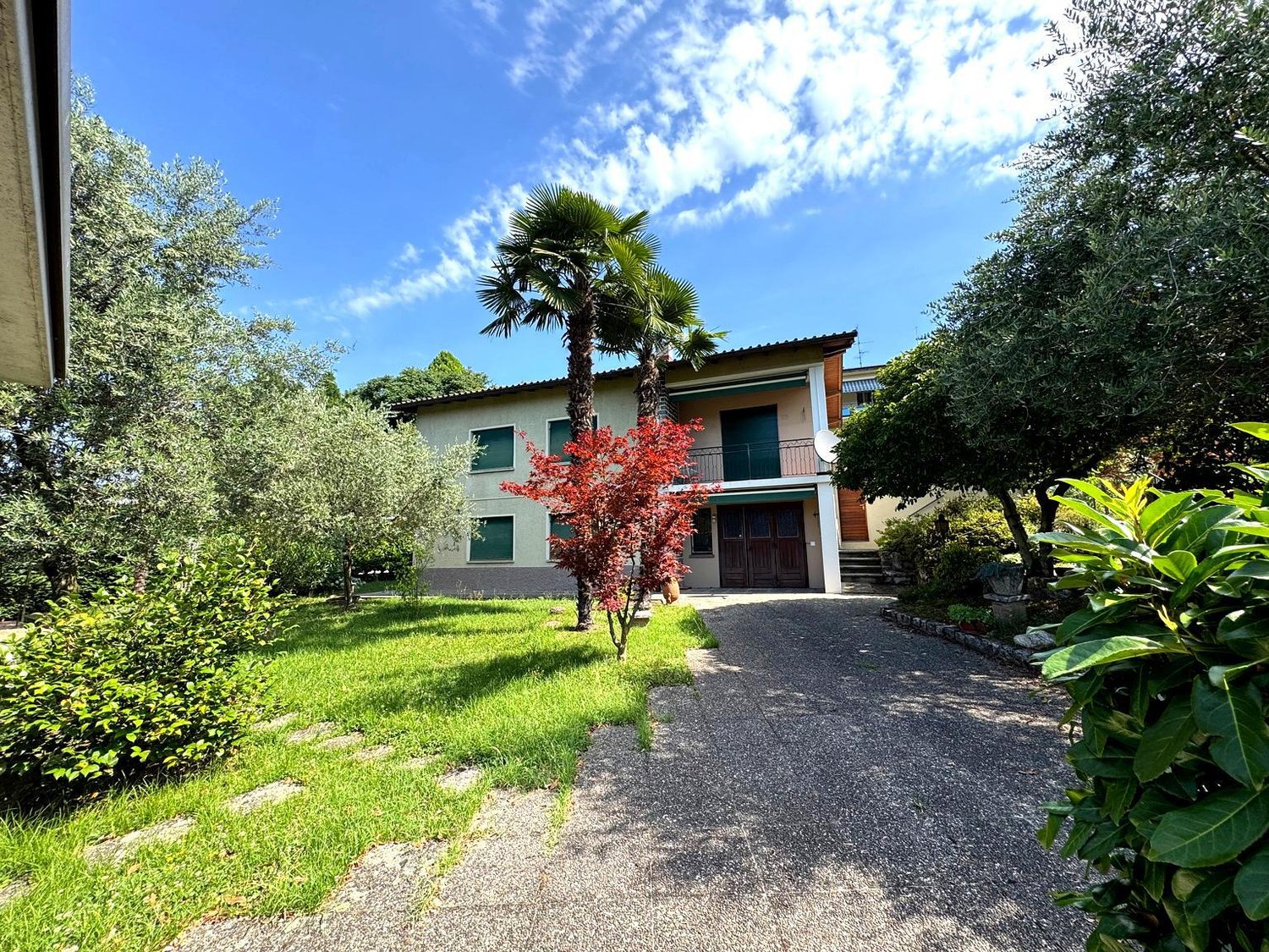 Two-story house with green exterior, large windows, balconies, surrounded by lush greenery including palm trees and other vegetation. Gravel driveway leading to the house.
