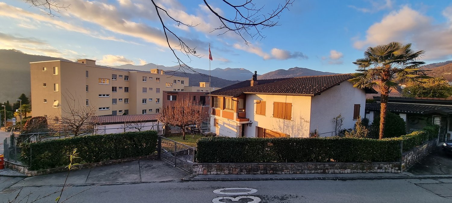 a single family house surrounded by hedges and greenery, a big building in the background, mountains in the distance