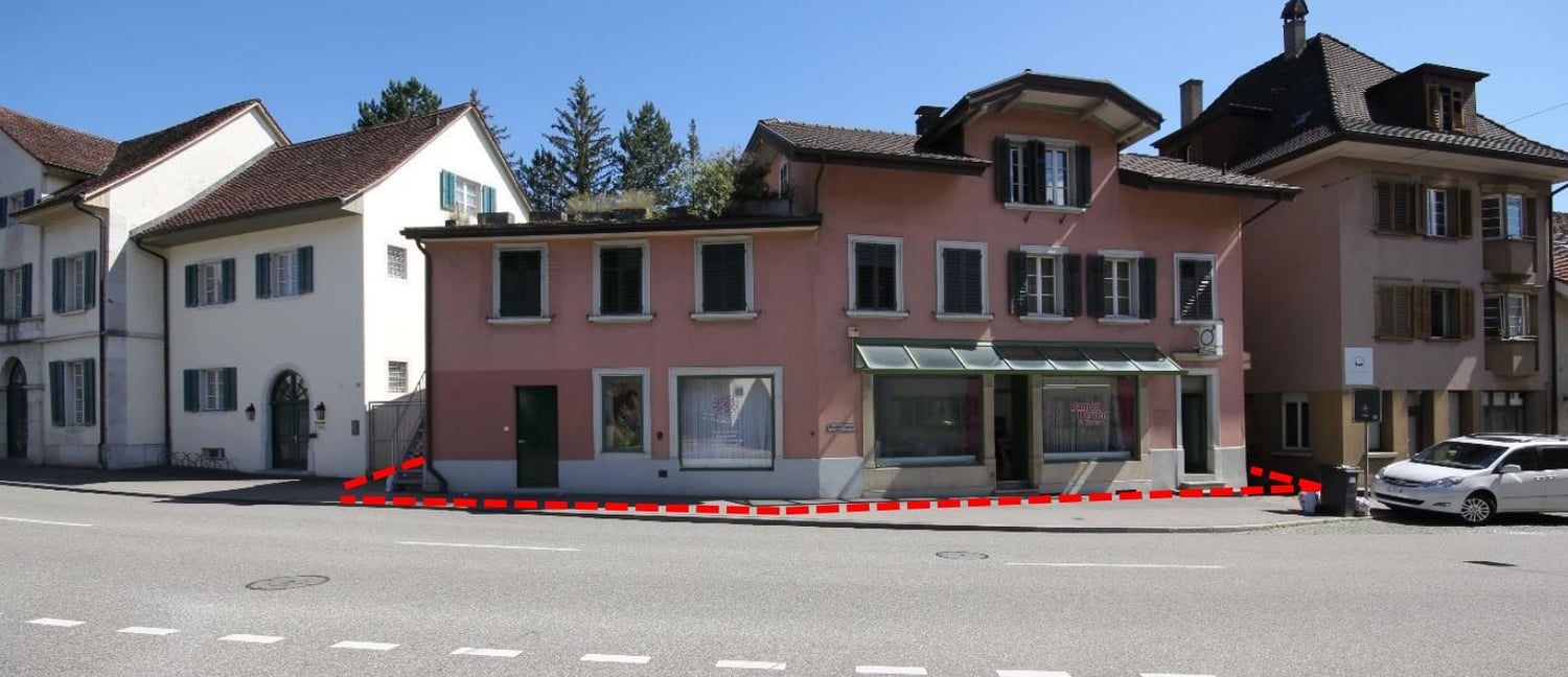 2-story pink and white buildings with slanted roofs, windows, and a storefront on the ground floor. There is a parking area in front of the buildings with a red and white barrier.