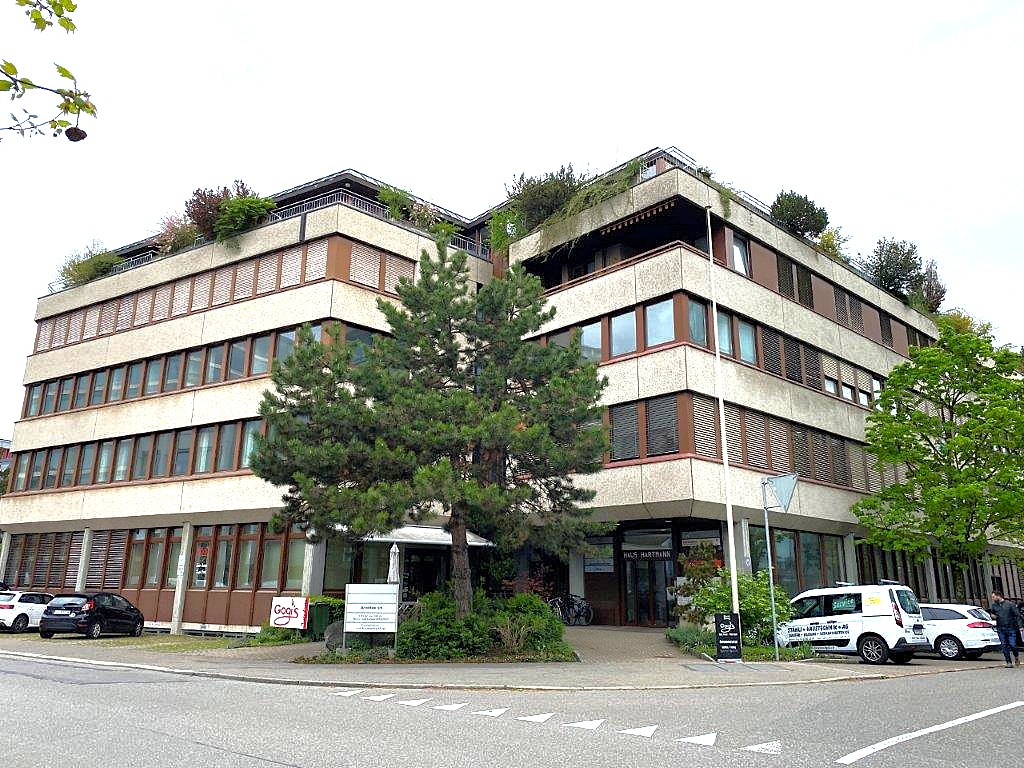 Multi-story office building with multiple windows, brown exterior, trees in front, and a roof garden.