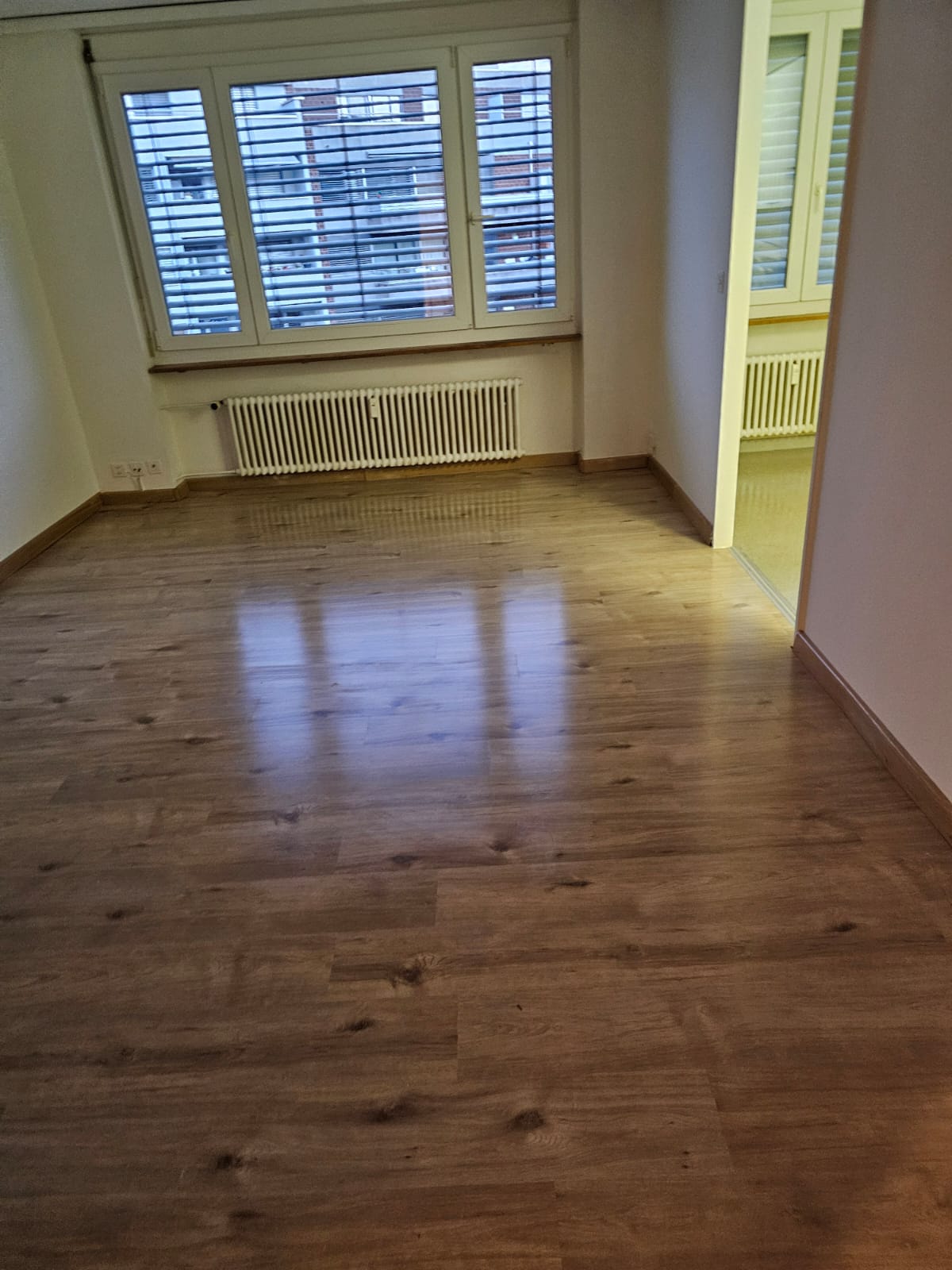 Empty room with white walls, white ceiling, wooden floor, large window with white shutters, white radiator