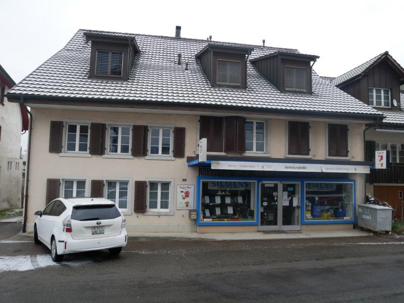 2 story house, gray roof, white windows, brown shutters, Siemens store on the ground floor, white car parked in front
