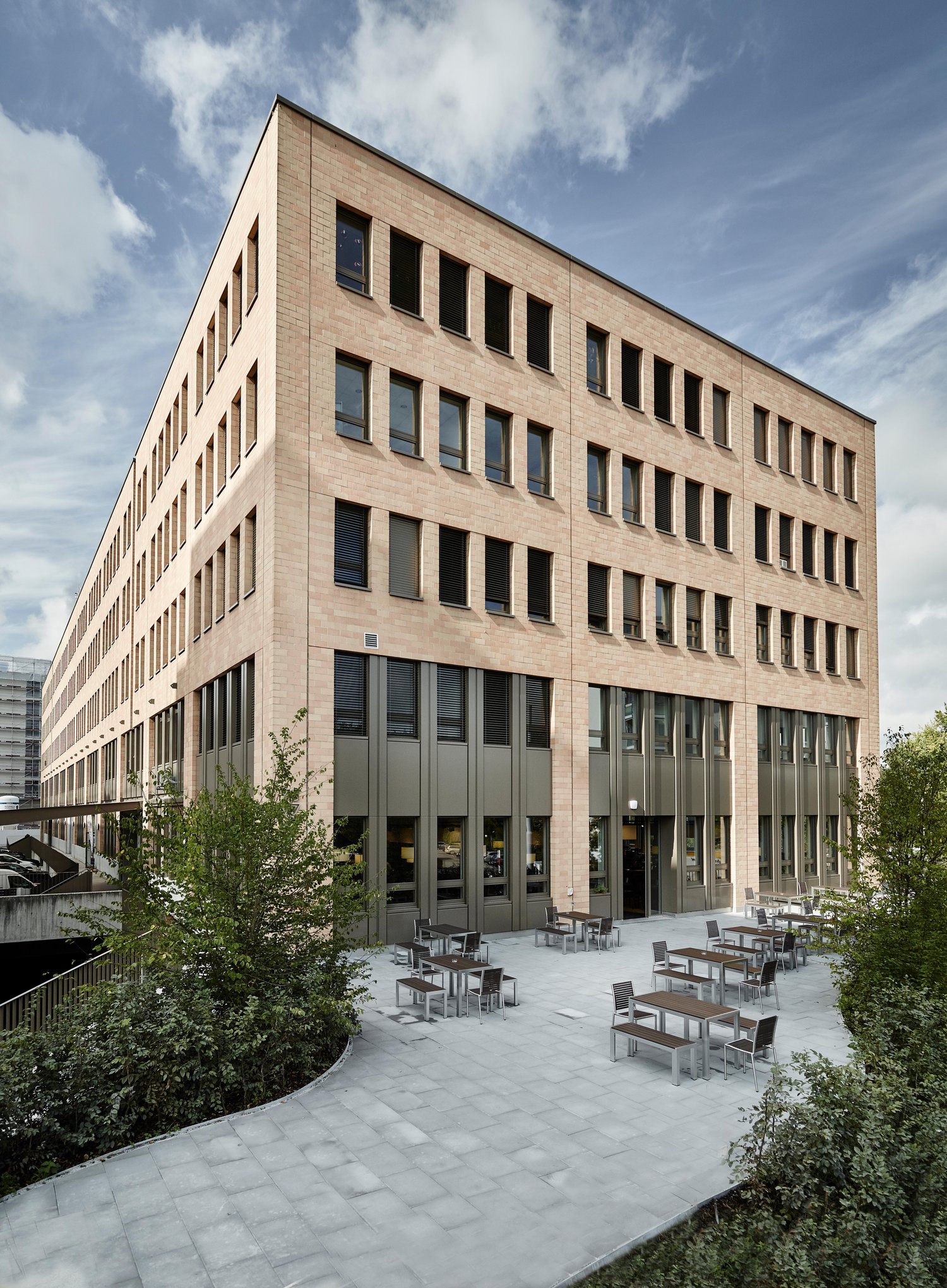 Multi-story office building with a modern, rectangular design featuring large windows and a paved outdoor seating area with tables and chairs.