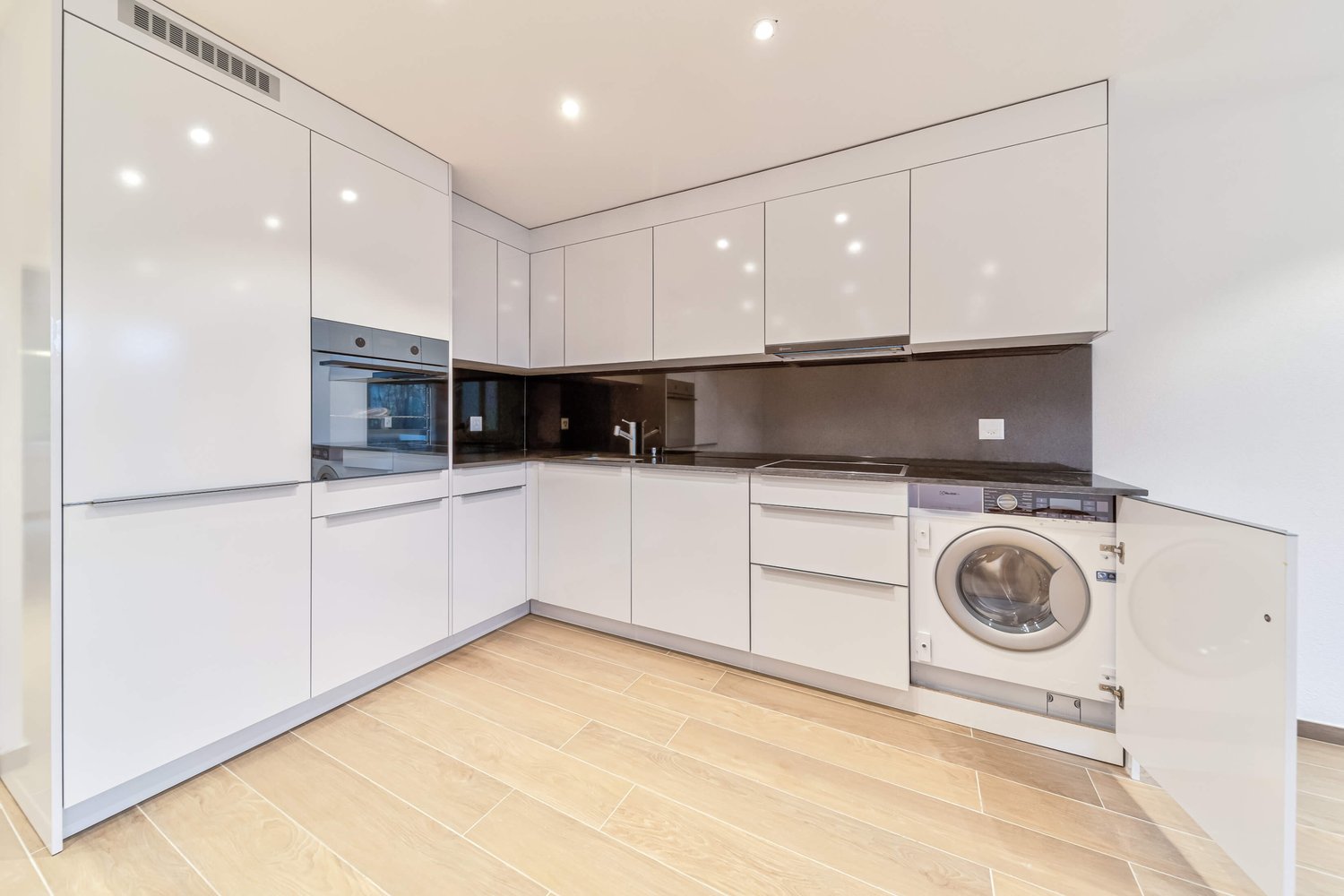 Modern kitchen with white cabinets, wooden floor, integrated dishwasher, washing machine, black countertops, stainless steel sink, built-in oven.