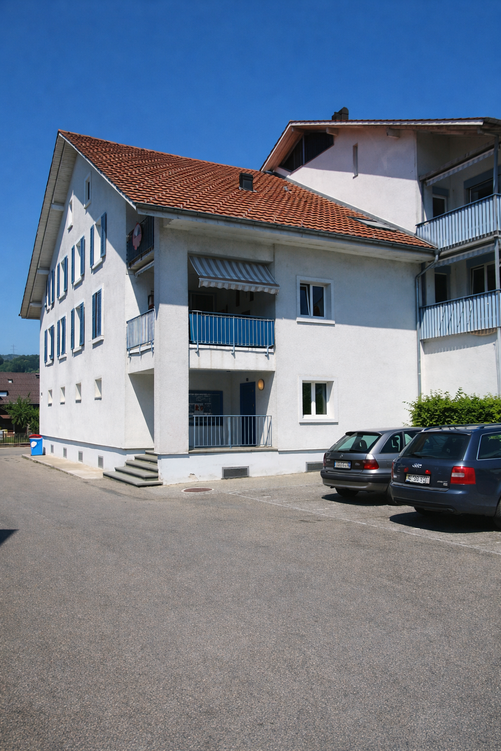 Two story house, white exterior, blue windows, brown tile roof, two balconies, car parked in front