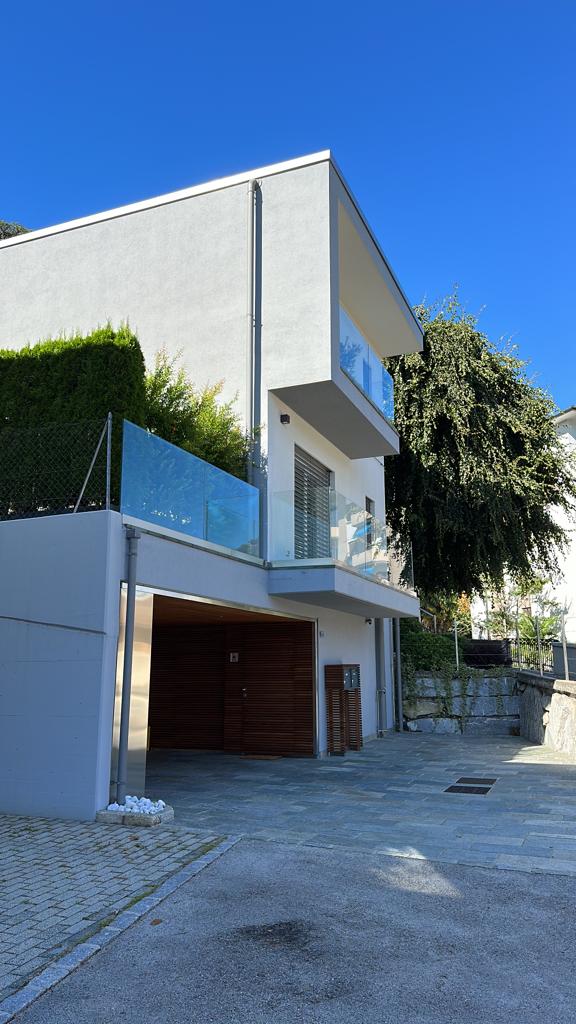 2-story modern house with a garage, balcony, and glass elements on the facade. The house has a minimalist, contemporary design with a flat roof and is surrounded by greenery.