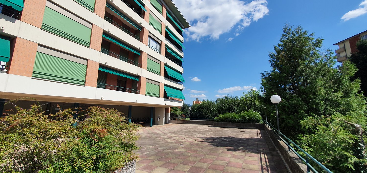 Large building with multiple floors, brick facade, multiple balconies, parking area, green awnings