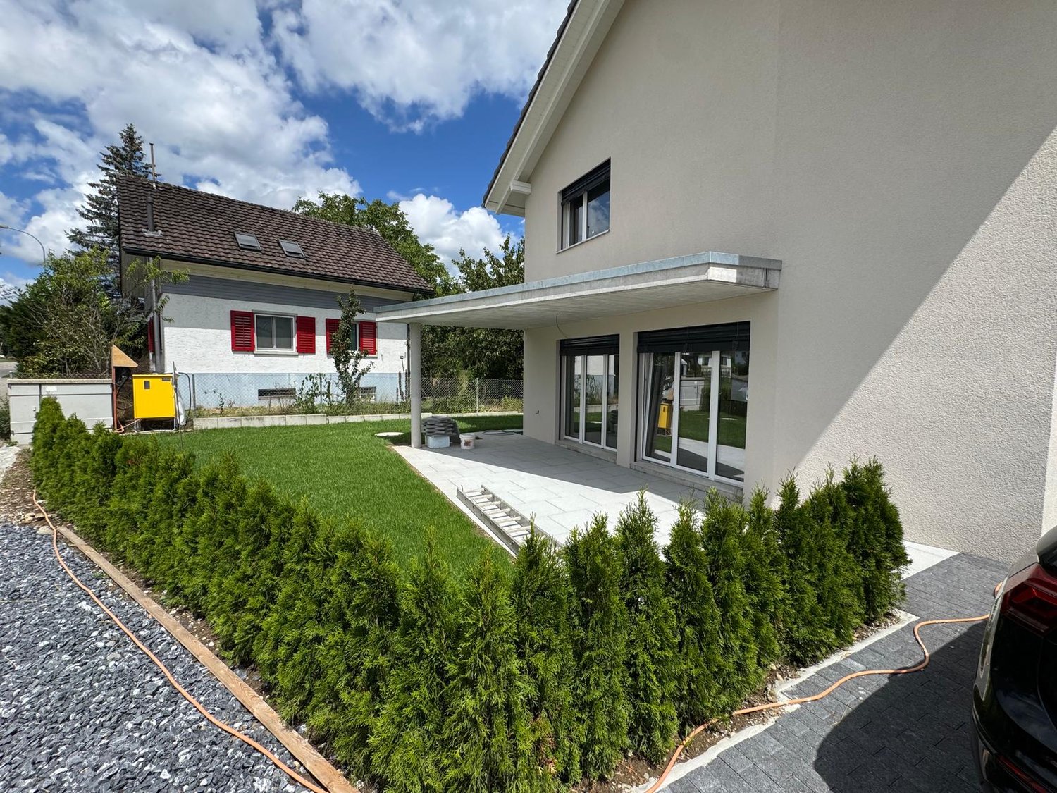 Modern single family house with a grassy front yard, glass doors, white walls, and a covered area for vehicles. It has another house in the back.