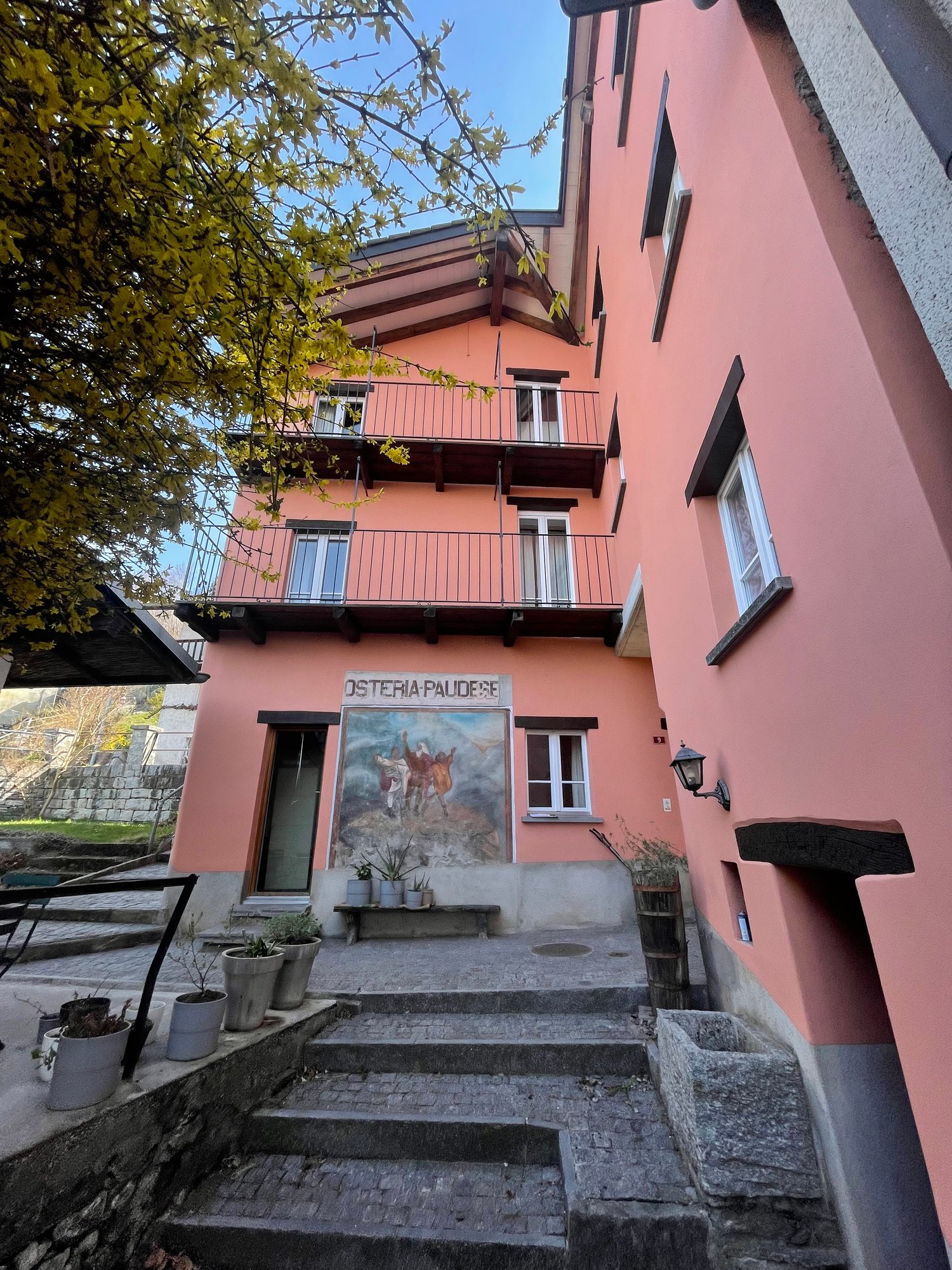 A two-story pink building with balconies, surrounded by trees. The building has a mural painting on the front and a set of stairs leading to the entrance.