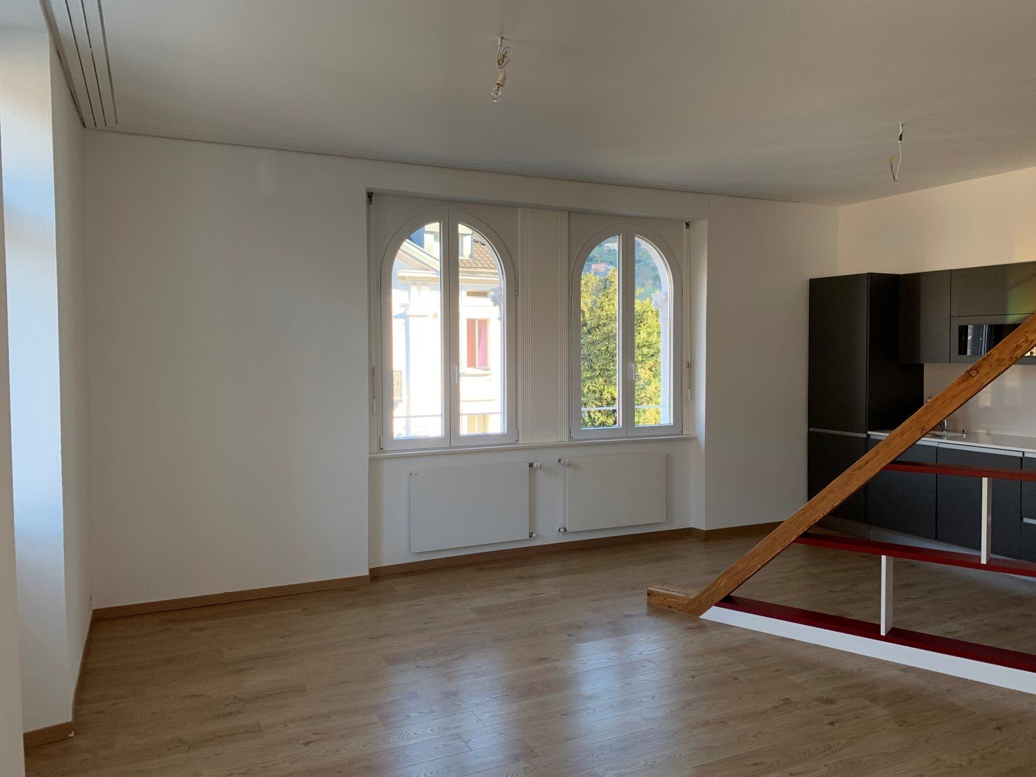 Spacious living room with large arched windows, hardwood floors, and a modern kitchen area with black cabinets.