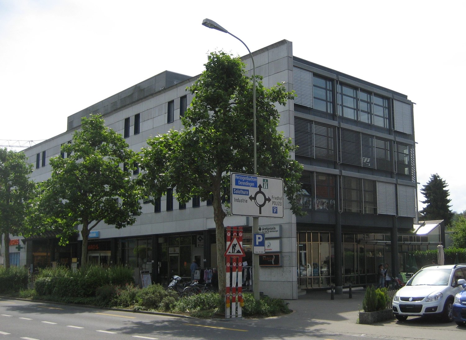 Multi-story office building, white exterior, glass windows, ground-level storefronts, trees and bushes in front, cars parked on street, street light in front.