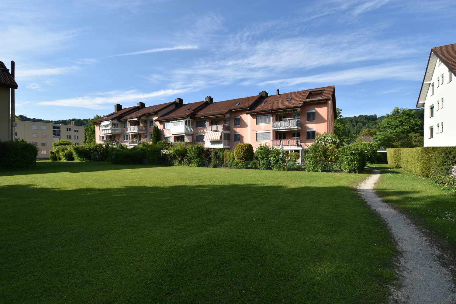 A row of apartment buildings with brown roofs and balconies, set against a backdrop of greenery and a wide grassy field.