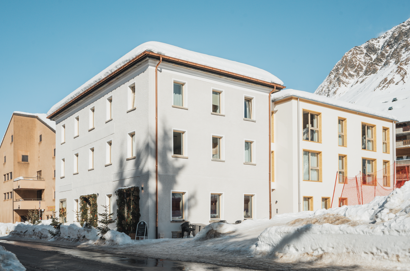 A multi-story white building with a sloped roof, surrounded by snow-covered mountains. The building has several windows and a balcony or terrace visible.