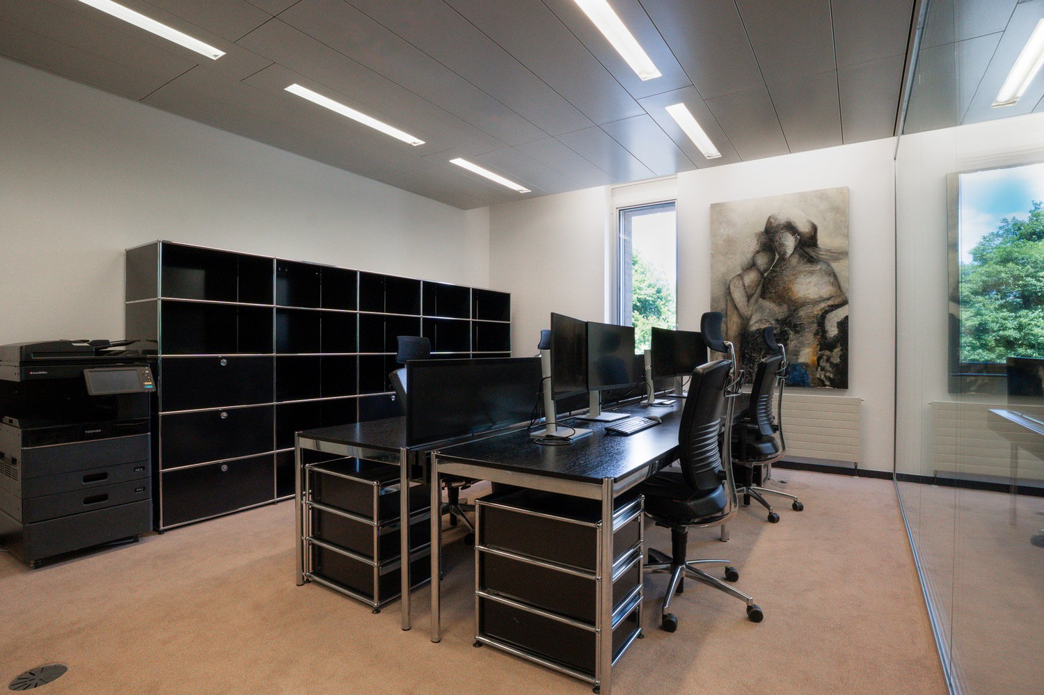 The image shows a modern office space with multiple workstations. The room has a sleek and minimalist design, with black desks, chairs, and storage units. There are several computer monitors and printers on the desks, indicating this is a workspace for of