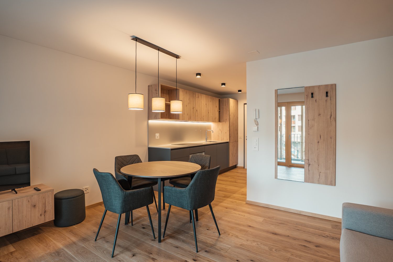 wood floor, round table, grey chairs, kitchen area, mirror, wooden door, TV, ottoman