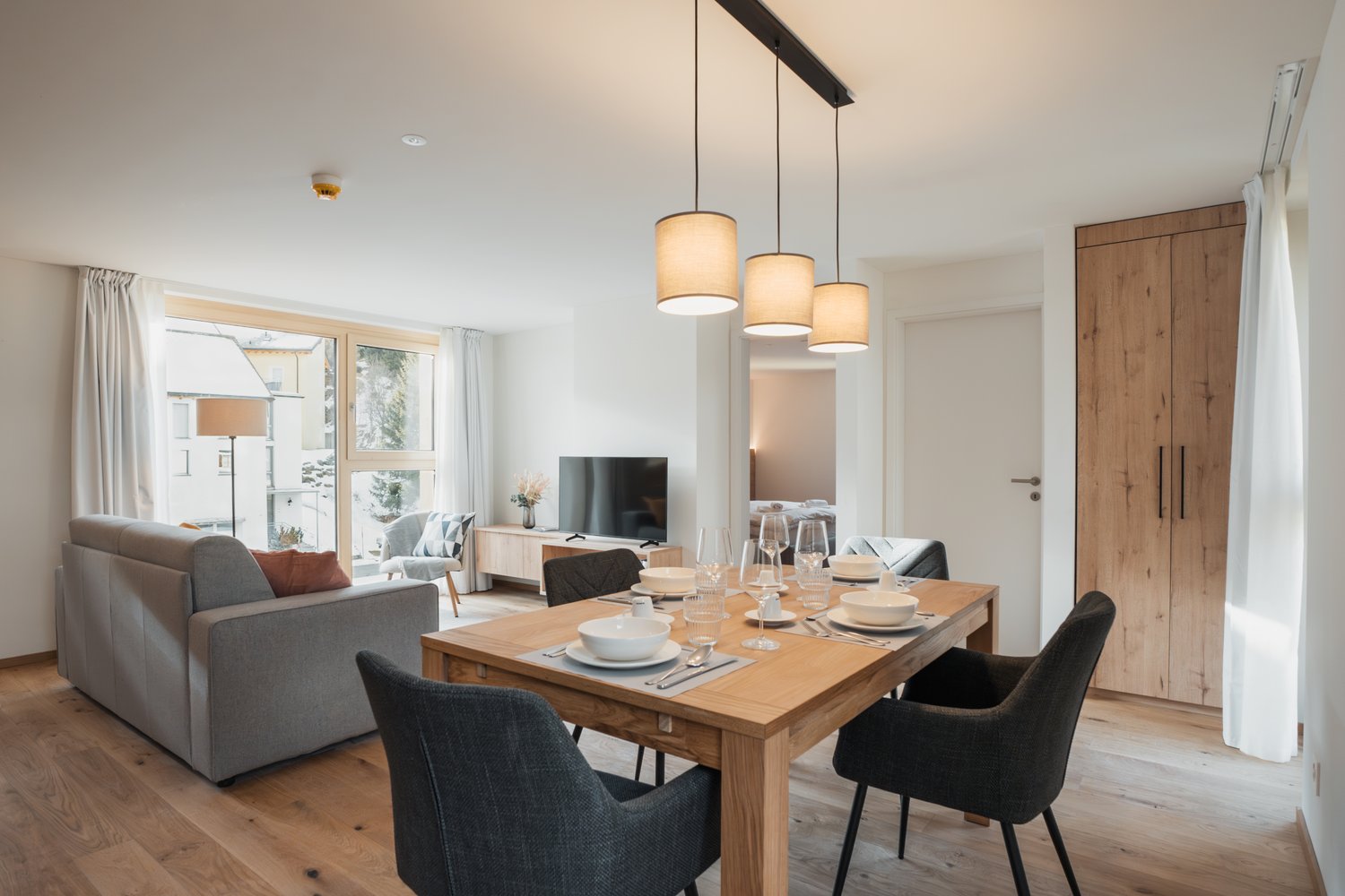 modern dining area with wooden table and chairs, gray sofa, table set for 6, large windows, tv, wooden cabinets