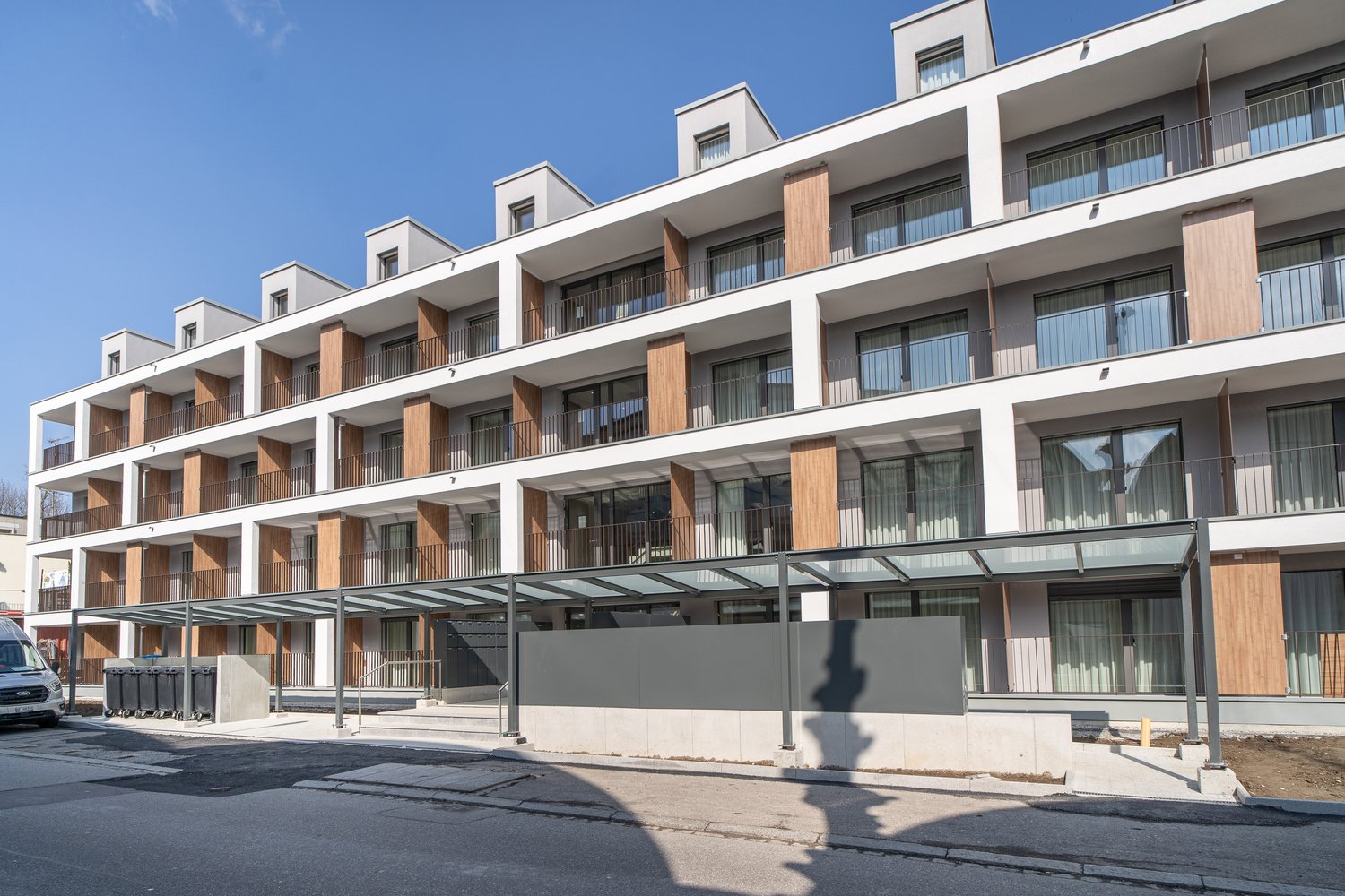 Multi-story apartment building with modern, minimalist design. The building features balconies on each floor, with a mix of glass and wood accents. There is a covered entrance area at the ground level.