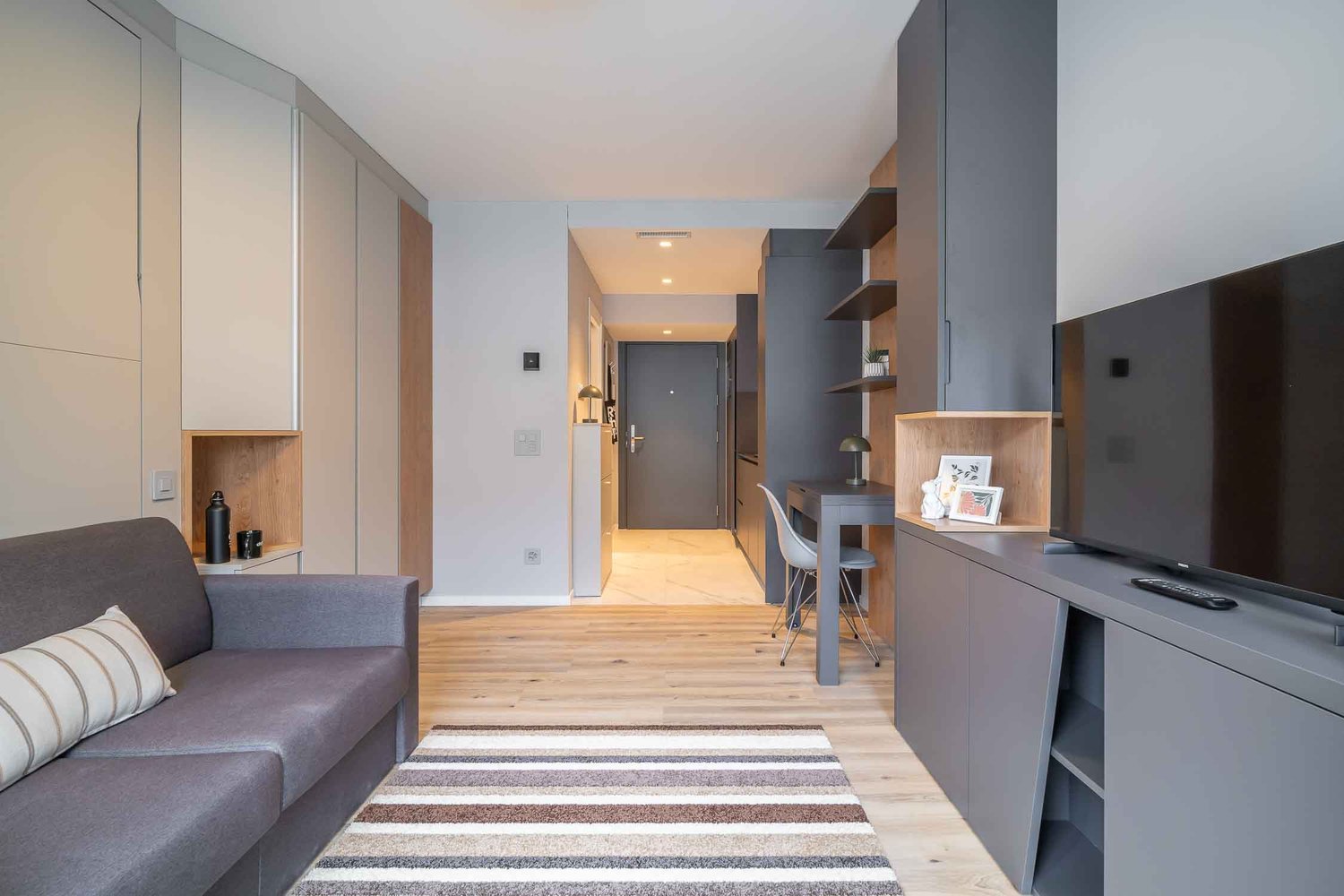 Open-plan studio apartment with living room, kitchenette, and dining area. Gray and wood tones throughout, with a gray sofa, wooden floors, and built-in shelving and storage. Entrance hallway leads to the apartment. Dishwasher visible in the kitchenette a