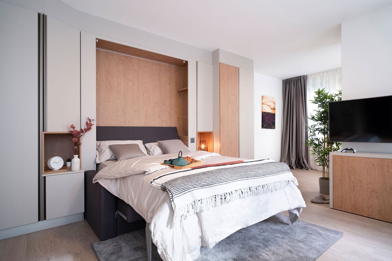 Spacious bedroom with wooden paneling, large bed, bedside tables, and a TV. The room has a cozy and modern feel with gray curtains and a rug on the floor.