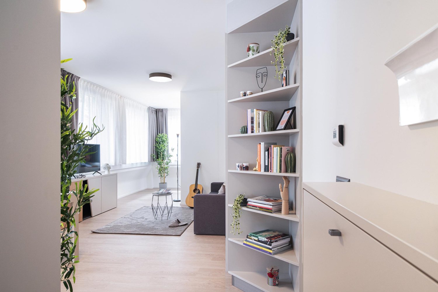 Spacious living room with white walls, hardwood floors, large windows, built-in shelving unit, potted plants, guitar, and a couch