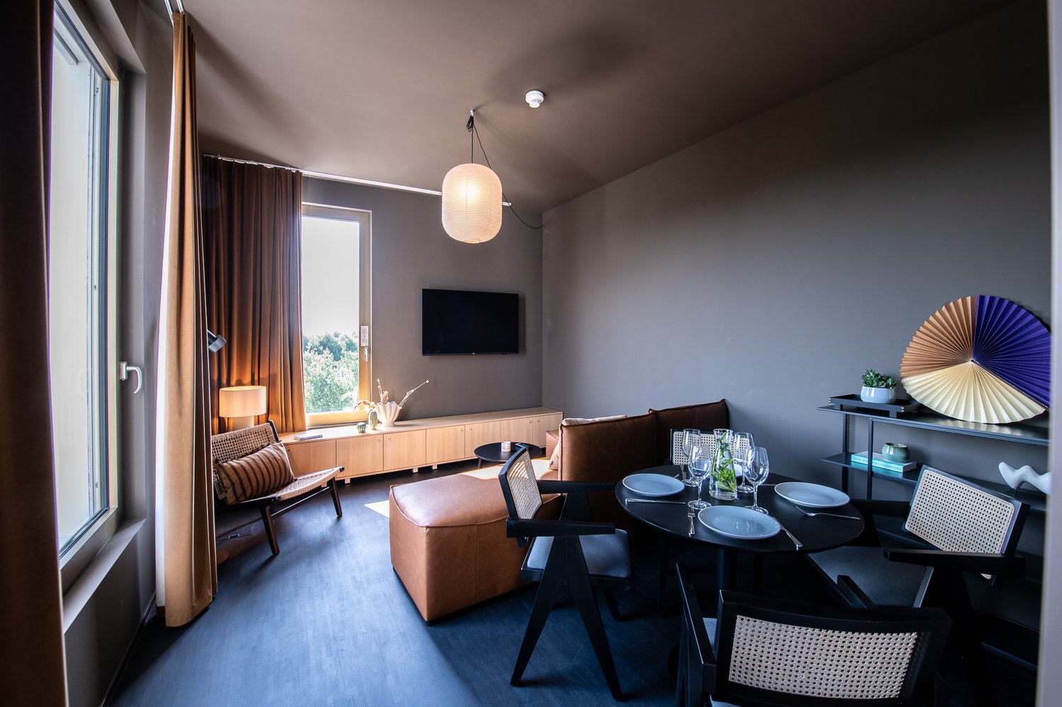 Spacious living room with large windows, brown curtains, wooden TV stand, leather sofa, dining table with chairs, and decorative elements like a fan-shaped wall art piece.