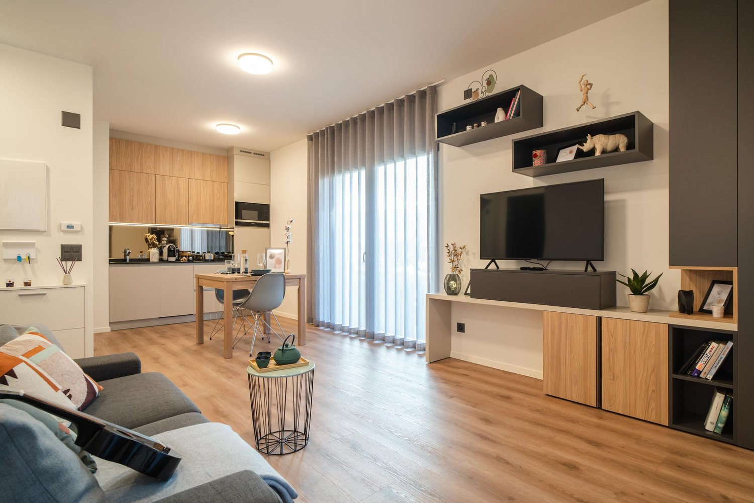 modern open plan living room, wooden floor, gray couch, wooden dining table, TV on entertainment unit, open kitchen with breakfast bar