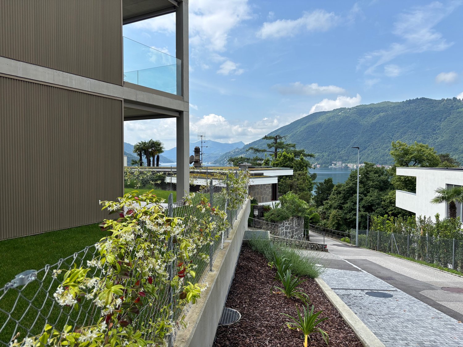 Modern building with large windows overlooking a scenic lake and mountains. The building has a balcony or terrace area with lush greenery and landscaping. The surrounding area features a paved walkway and additional landscaping elements.