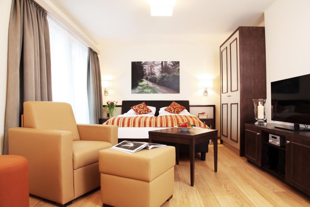 Spacious bedroom with hardwood flooring, large windows, a king-size bed with striped bedding, a seating area with two yellow armchairs and ottomans, a TV stand, and a framed artwork on the wall.