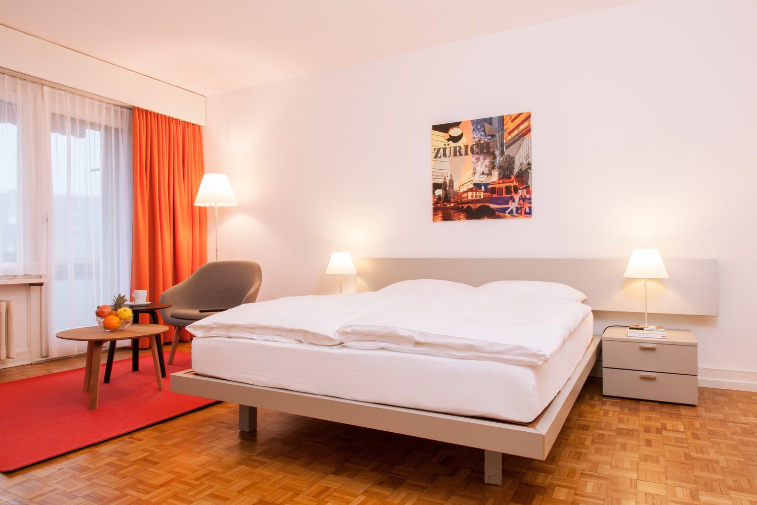 Spacious bedroom with a large bed, wooden floor, orange curtains, side tables with lamps, and a framed artwork on the wall depicting a city scene.