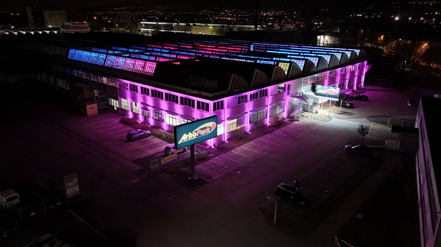 Large commercial building with colorful LED lighting on the exterior, parking lot in the foreground, and a sign for 'Arbor Park' visible.