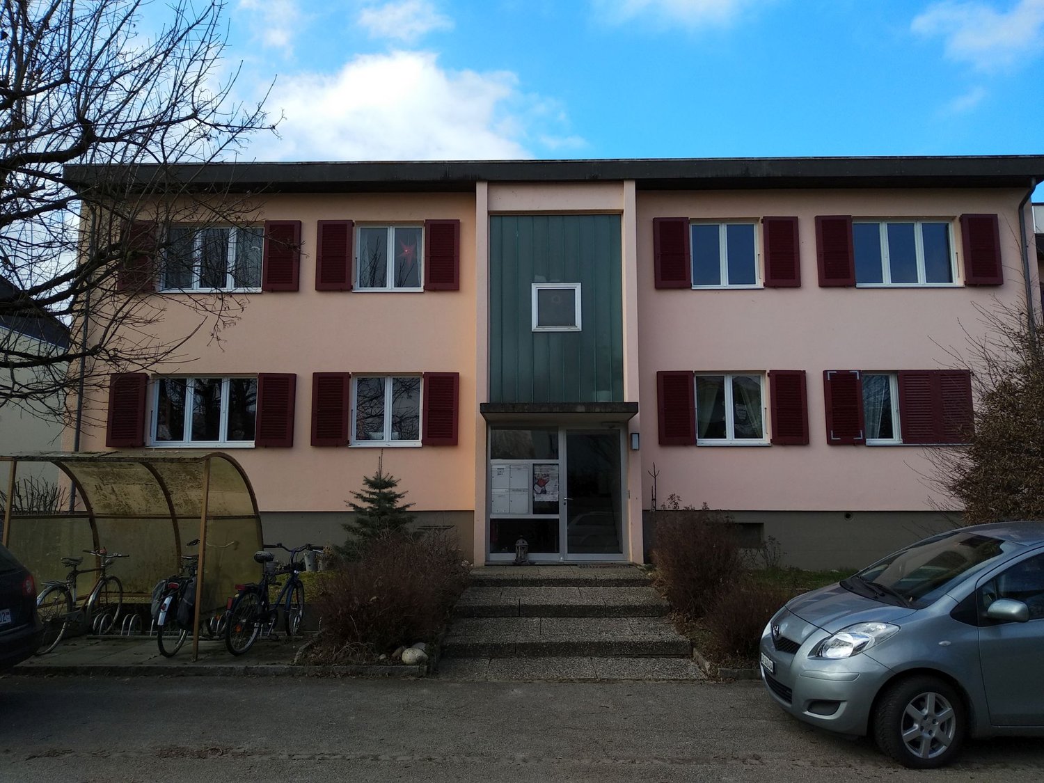 Multi-unit residential building with 4 floors, red shutters, entrance with a small staircase, bicycles parked in front