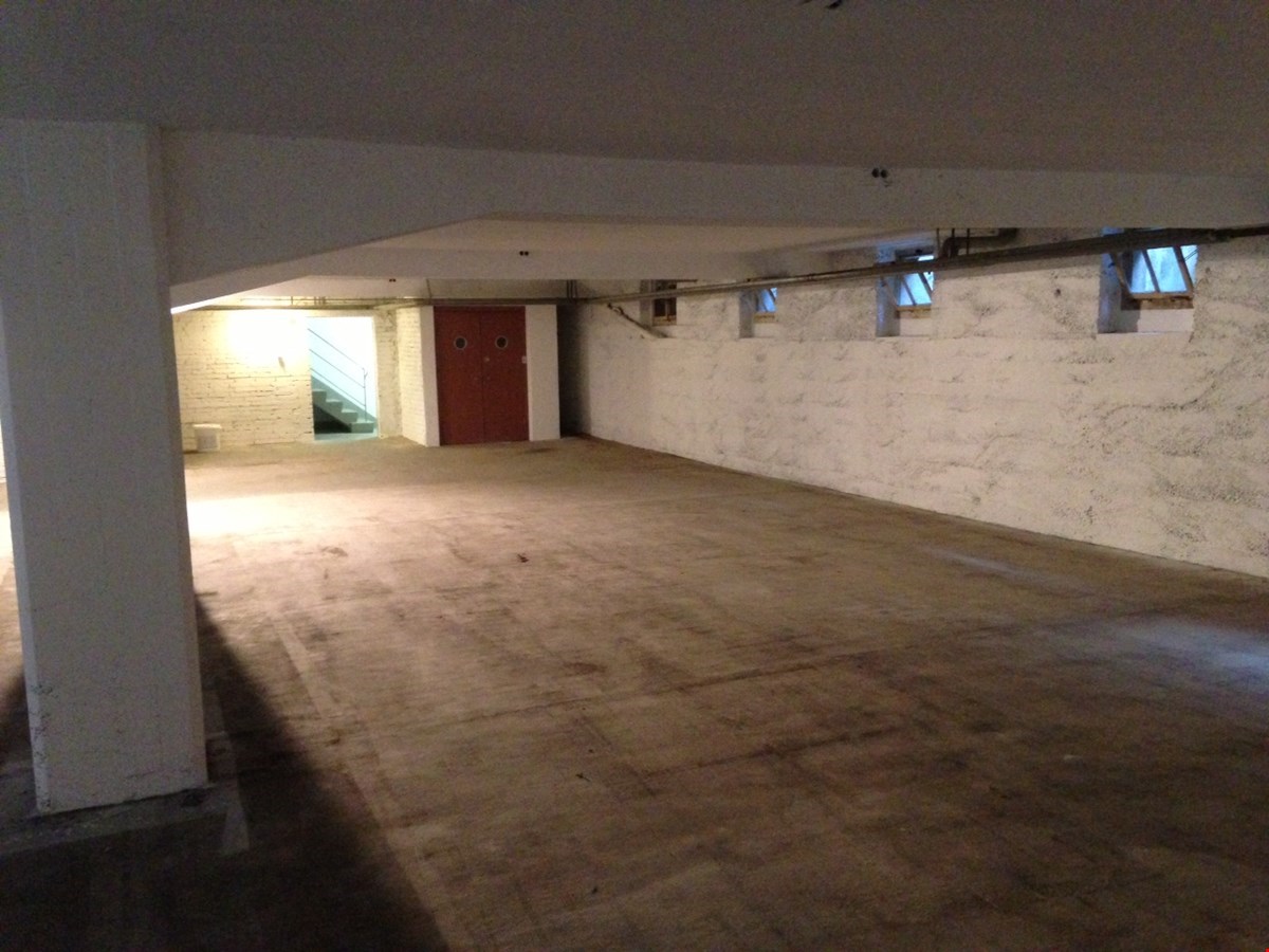 Empty room with concrete floor, white walls, red door, staircase to the left, three windows on the wall, light pipe coming from the ceiling.