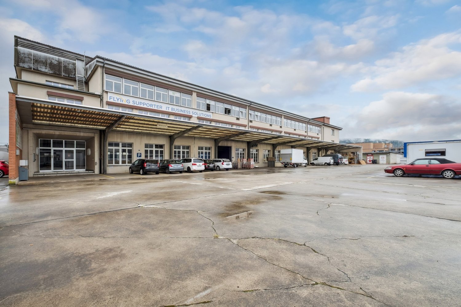 Industrial building with multiple windows, large parking area, cars parked in front, sign reading 'FLYING SUPPORTER IT BUSINESS SOLUTIONS'.
